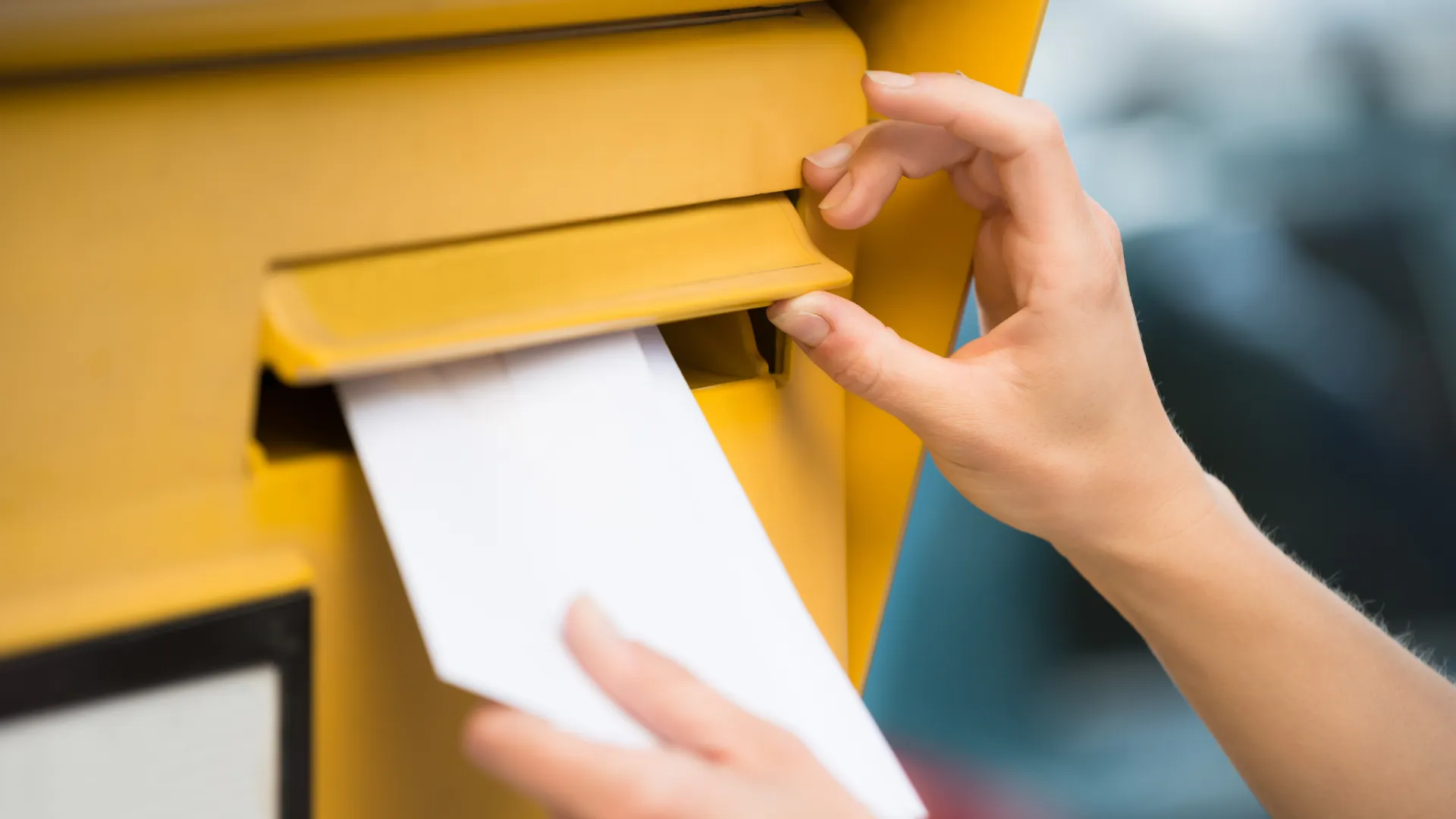 weibliche Hand steckt einen Briefumschlag in einen gelben Postbriefkasten